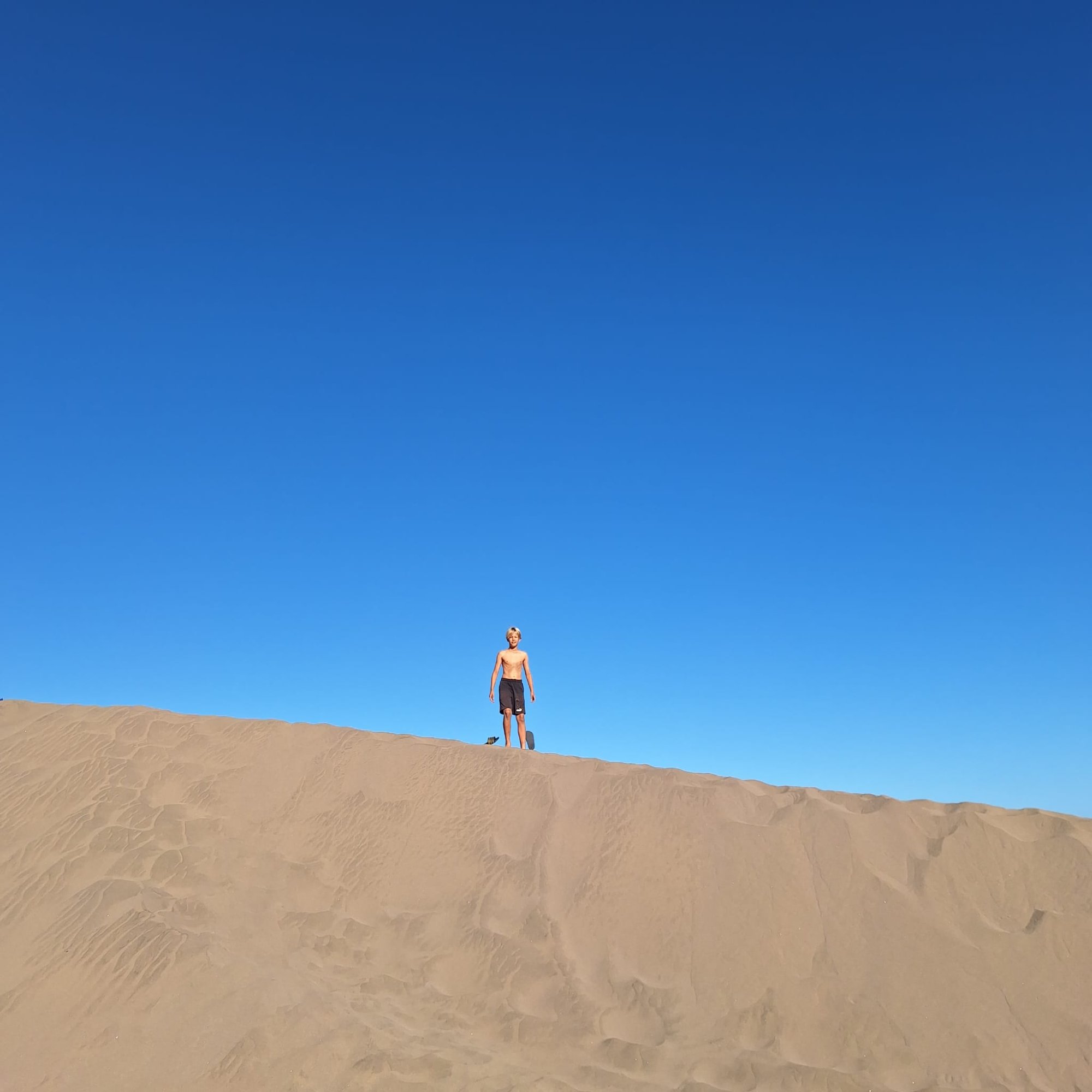 Young boy standing alone on top of a high sand dune against a deep blue sky