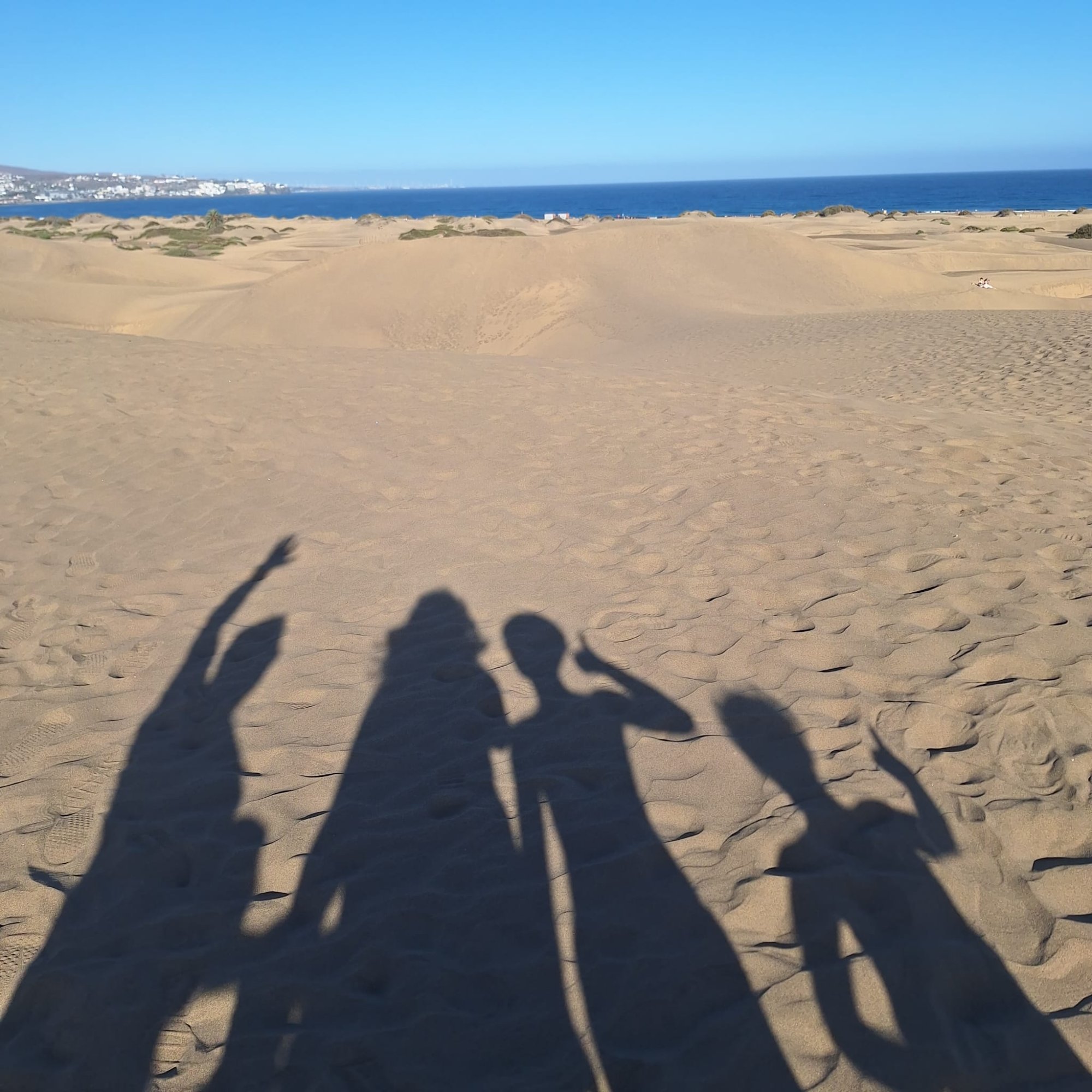 Family shadows cast across the golden sand dunes with the ocean and Playa del Inglés visible in the distance