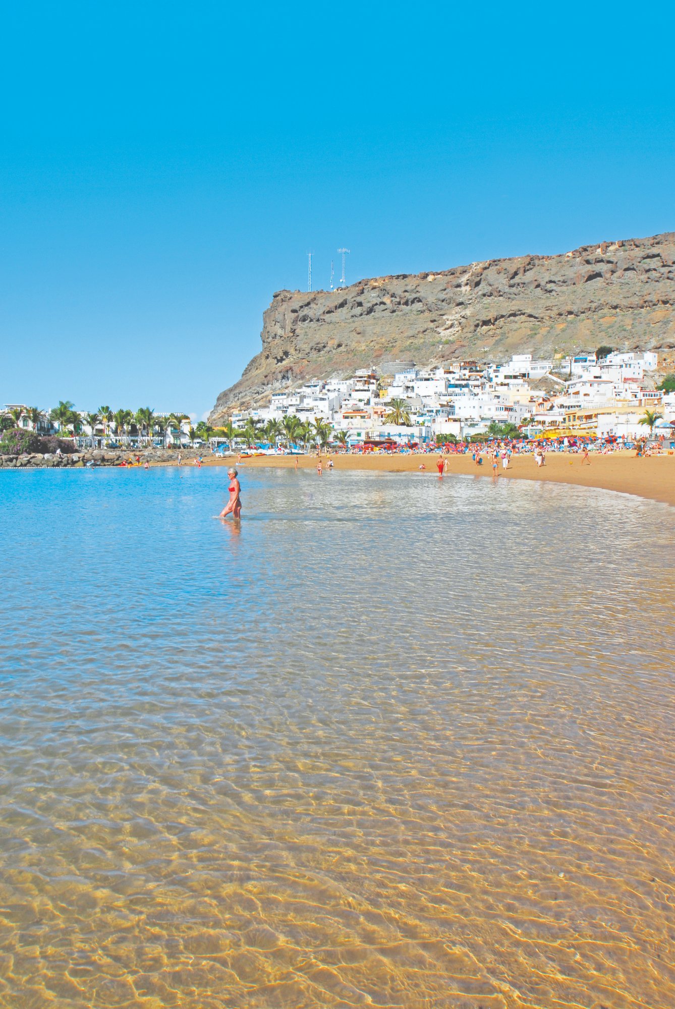 Crystal clear shallow water at Puerto de Mogán beach with colourful village and cliffs behind